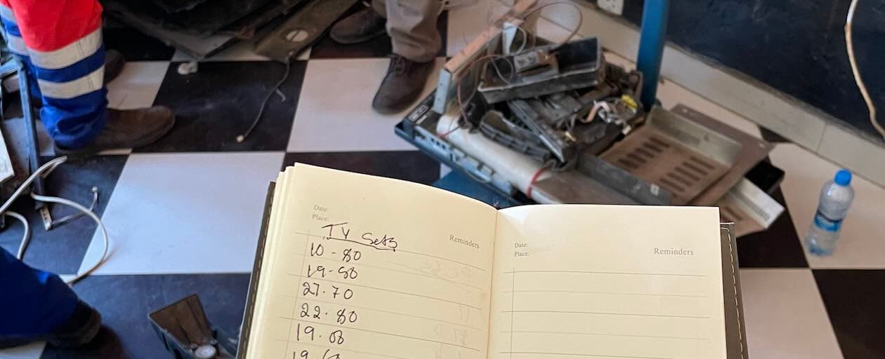 Open notebook containing handwritten inventory lists for TV sets held over a black and white checkered floor in an electronics workshop