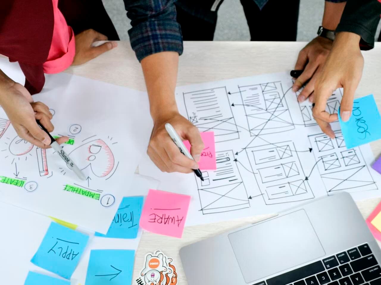 A desk with a MacBook, sticky notes, and paper, with multiple pairs of hands sketching UX wireframes and illustrations on the paper sheets