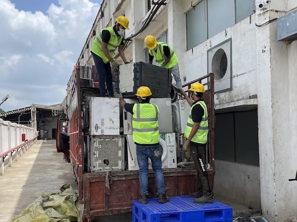 Four workers in safety vests wearing helmets unload old washing machines from the back of a truck