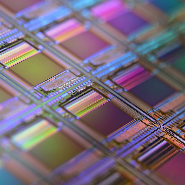 Macro view of a silicon wafer featuring a grid of rectangular semiconductor chips reflecting iridescent purple and blue colors